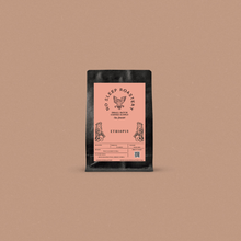 Load image into Gallery viewer, Fresh roasted coffee in a black bag with a pink label displayed on a light brown background