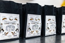 Load image into Gallery viewer, A row of black coffee bags with white labels reading Ambition Blend on a reflective countertop.