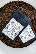 Load image into Gallery viewer, Two black coffee bags labeled Ambition Blend in a white bucket resting on a bed of fresh roasted coffee beans.