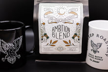 Load image into Gallery viewer, A bag of Ambition Blend coffee from No Sleep Roastery is centered between two mugs on a dark surface. The bag has a white illustrated label with owl graphics.