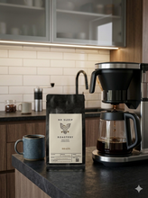 Load image into Gallery viewer, A bag of No Sleep Roastery whole bean Brazil fresh roasted coffee stands on a dark kitchen countertop next to a blue ceramic mug and a black coffee maker with brewed coffee. The background shows a white subway tile backsplash and wooden cabinets.