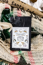 Load image into Gallery viewer, A black coffee bag labeled Ambition Blend by No Sleep Roastery, resting on a rustic background of burlap sacks.
